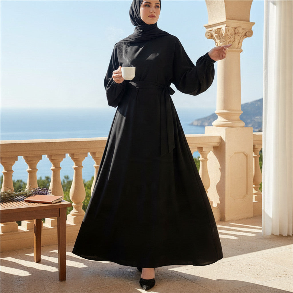 Woman wearing a black BelleNoor Nida fabric abaya with a matching hijab, standing on a balcony overlooking the sea.