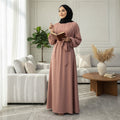 Woman wearing a dusty rose BelleNoor Nida abaya with a black hijab, standing in a bright, modern living room.